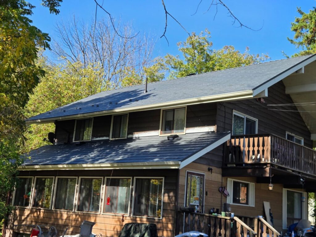 A two-story brown wooden house with a blue-grey metal roof, surrounded by lush trees under a clear blue sky.