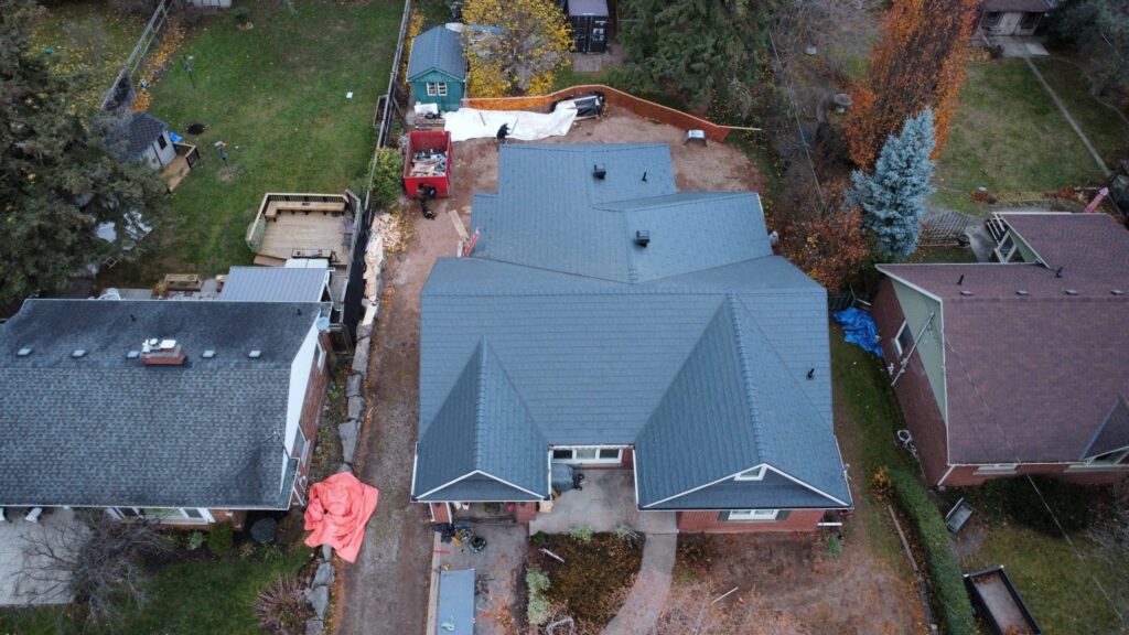 Aerial view of a neighbourhood featuring three houses. The central house has a dark grey, multi-faceted metal roof. Surrounding trees show autumn colours, with a red truck parked in the backyard.