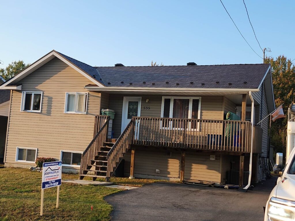 Single-story beige house with a wooden deck and stairs. A driveway leads to the entrance. A "Roof Pros Plus" sign is on the front lawn.