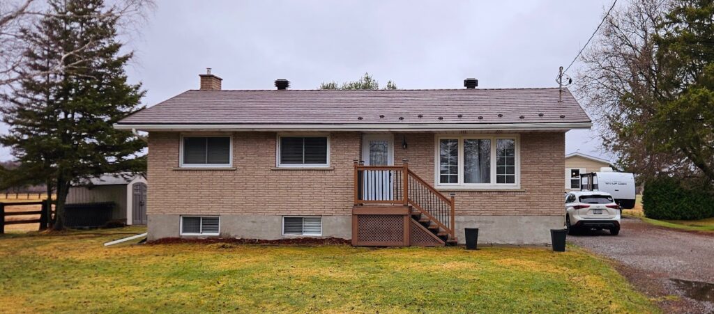 A brick house with a small wooden porch and stairs is surrounded by a grassy yard. 