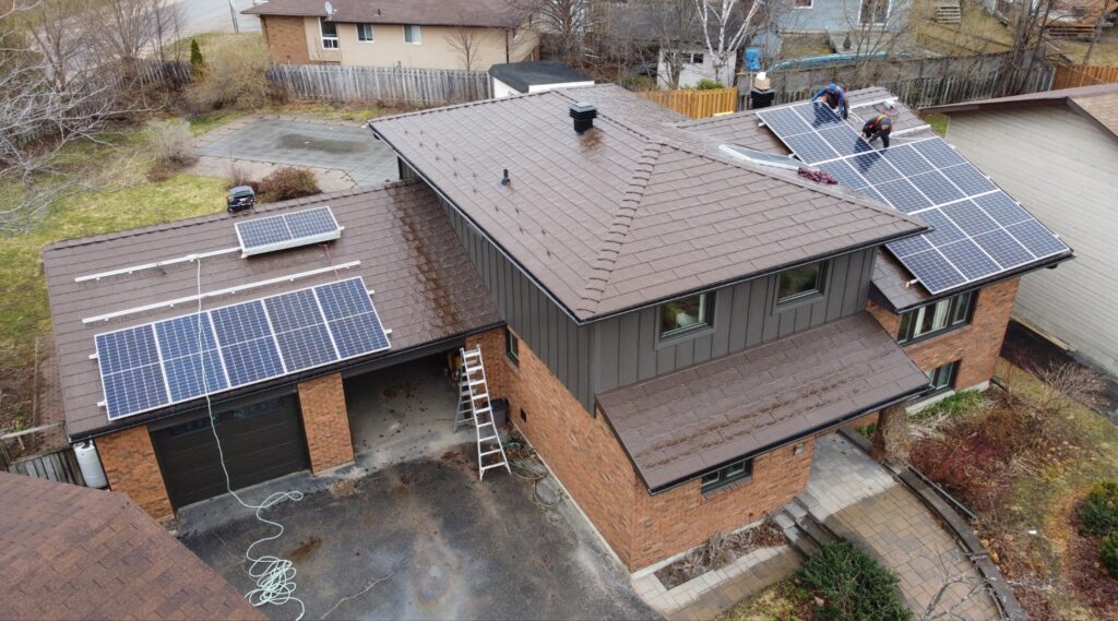 An aerial view of a modern brick house with solar panels being installed on the metal roof. Two workers are securing panels, surrounded by early spring vegetation.