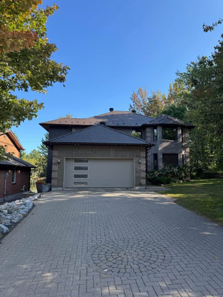 A two-story brick house with a grey garage door and dark metal roof, surrounded by lush greenery and trees under a clear blue sky. 