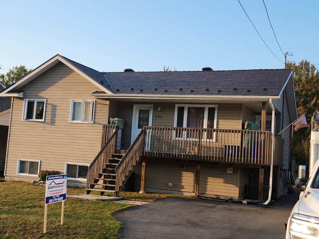  A brown house with white trim, an elevated wooden porch, stairs and a dark grey metal roof. A "Roof Pro Plus" sign is on the lawn. 