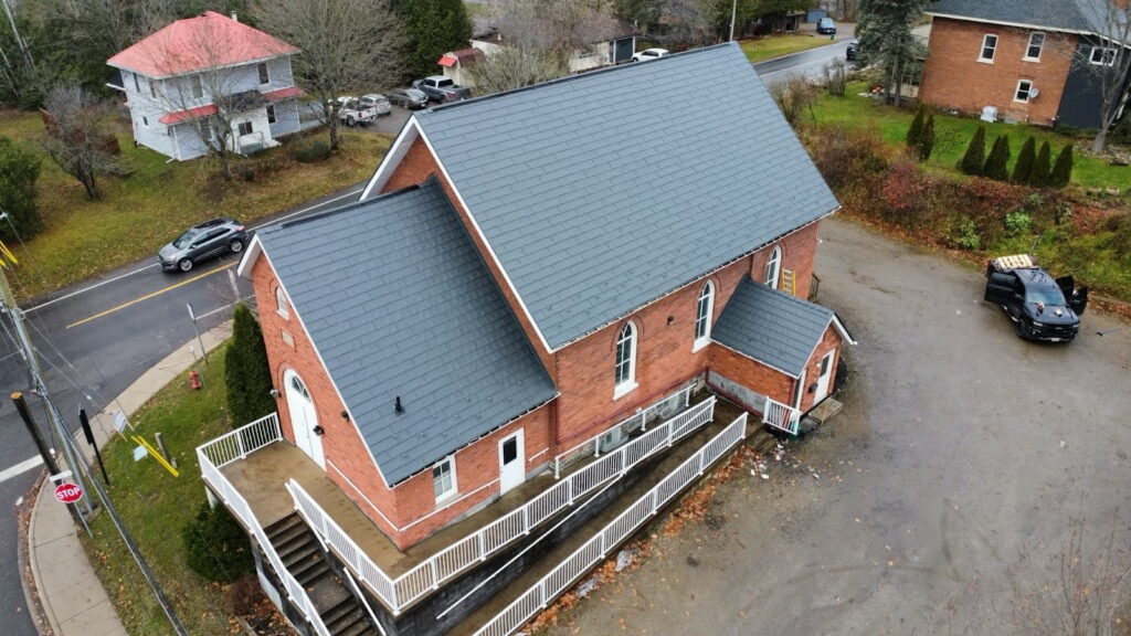 Aerial view of a brick building with a steep dark metal roof, adjacent white railings, and surrounded by autumn trees.