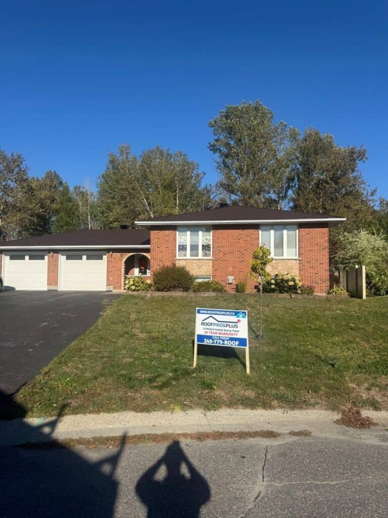 A brick house with two large windows, a dark metal roof, and a garage is under a clear blue sky. A sign for "RoofProsPlus" is on the front lawn.