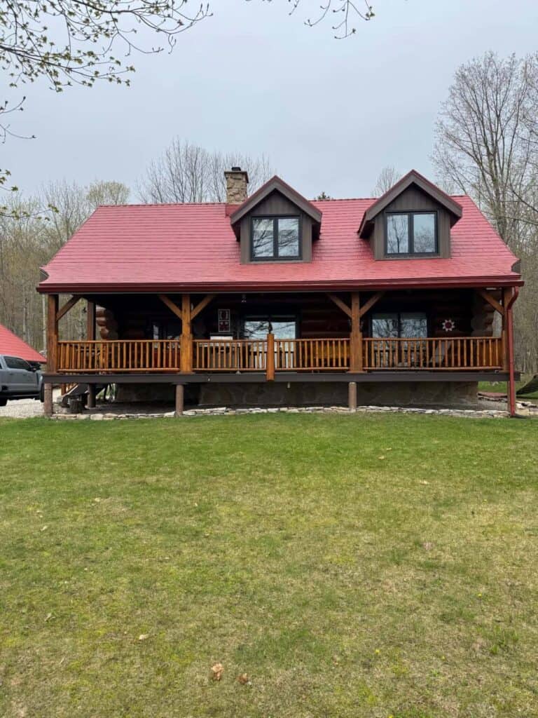 A rustic-style home with a red metal roof stands in a grassy yard surrounded by bare trees. 