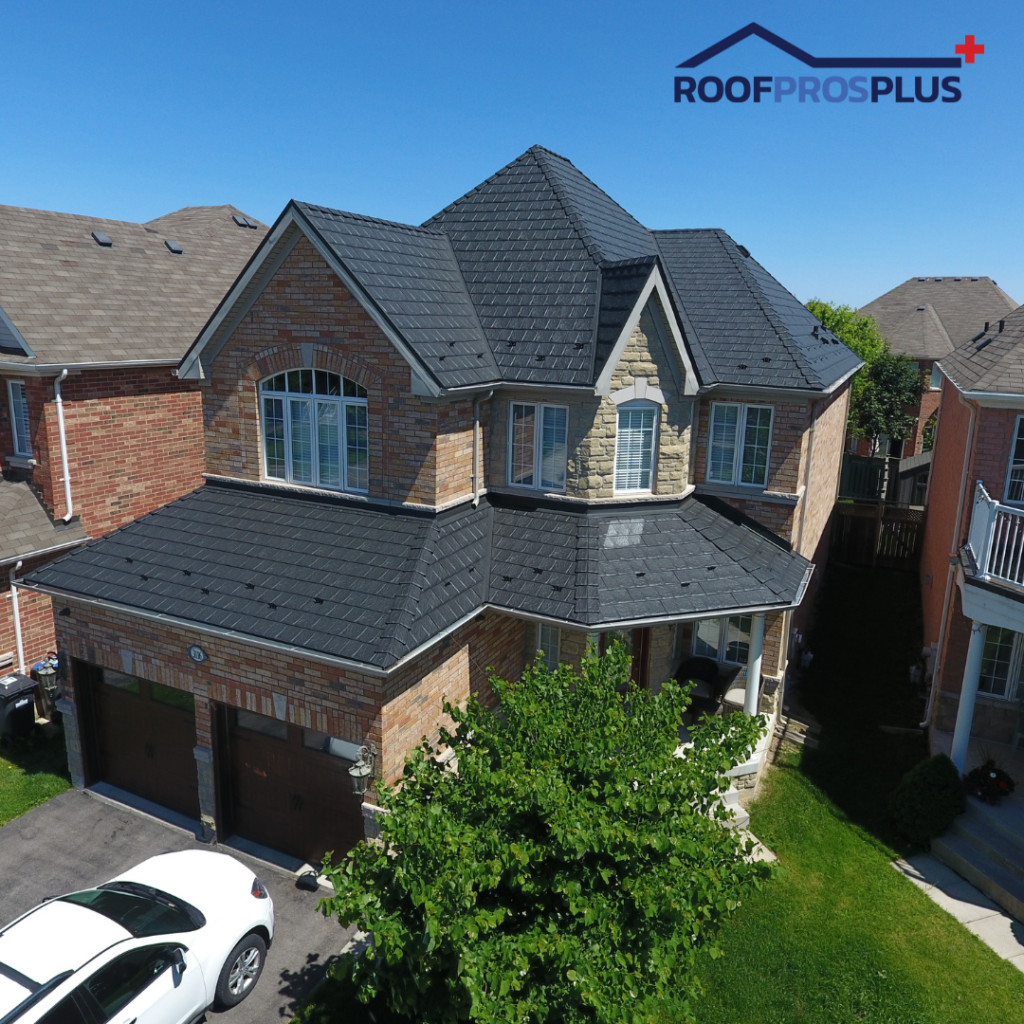 An aerial view of a two-story, brick house with a dark, metal roof under a clear blue sky. A white car is parked in the driveway. The Roof Pros Plus logo is in the top right corner.
