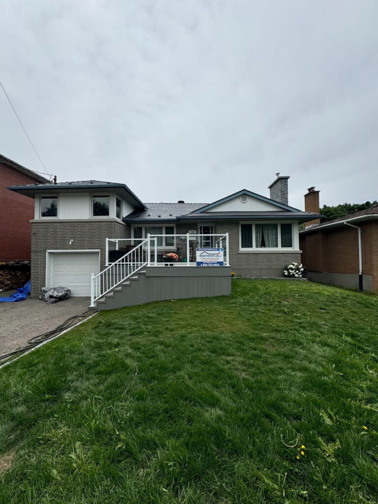 A two-story house with a gray brick facade, metal roof and a white railing porch sits under a cloudy sky. The front yard is grassy, and a "For Sale" sign is displayed hanging off of the porch.
