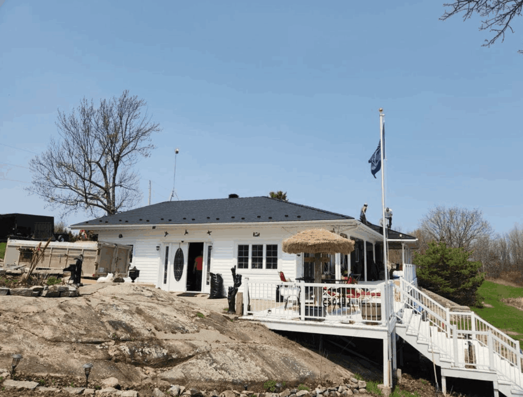 A single-story white house with a dark metal roof is surrounded by brown grass and leafless trees. A large deck with furniture and an umbrella is in front.