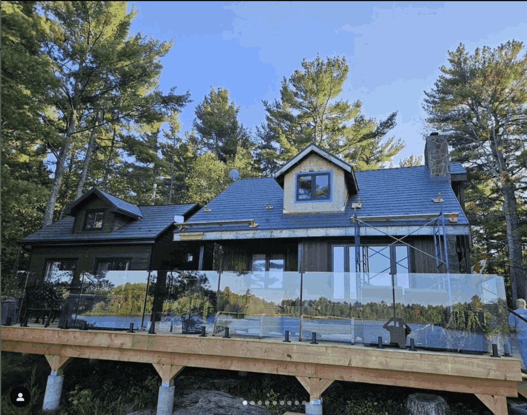 A rustic cabin with a dark roof under construction, surrounded by tall pine trees. It features a wooden deck with glass railings, set against a clear blue sky.