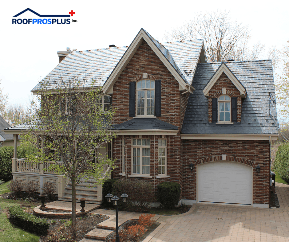 A two-story brick house with a steep grey metal roof, white-framed windows, and a small front porch. A leafless tree and shrubs are in the front yard. The Roof Pros Plus logo is in the top left corner of the graphic.