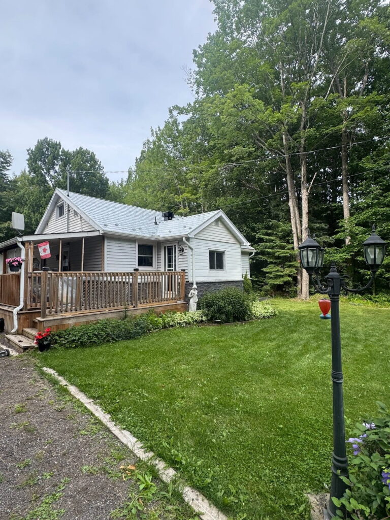 A cozy white house with a wooden porch and a metal roof surrounded by lush green grass and tall trees. A lamppost and hanging flower basket add charm. The picture is set under a cloudy sky.