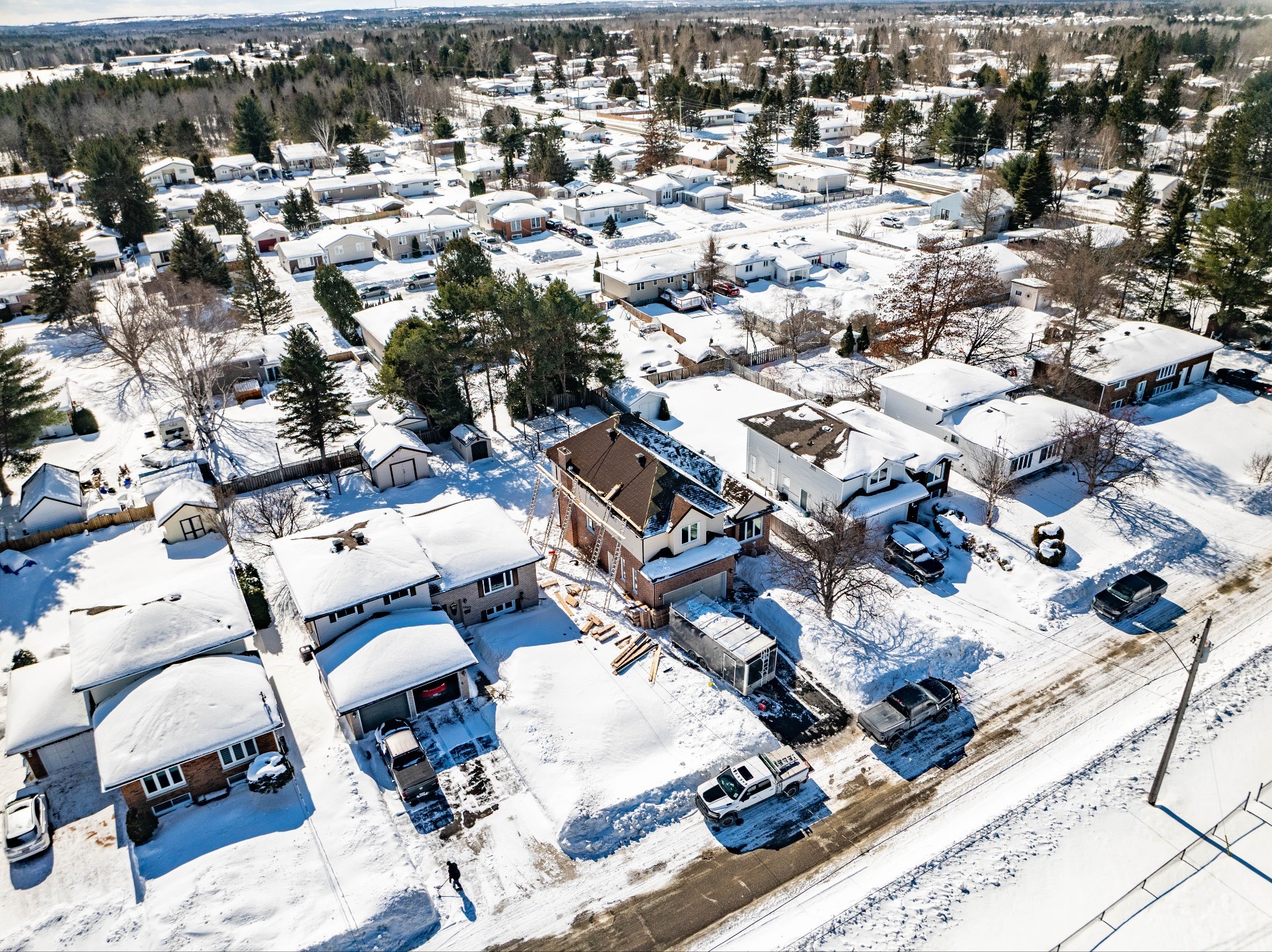 A snowy residential neighbourhood with snow-covered metal roofs and streets. Central house are under construction. Trees and parked cars line the road.