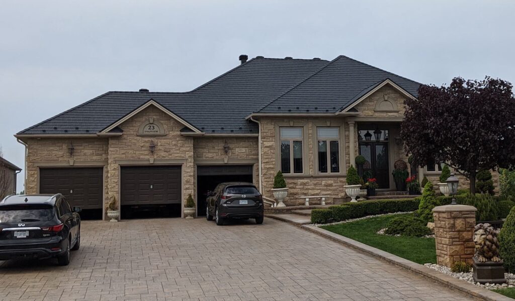 An elegant stone house with a dark metal roof, three garages, and a paved driveway. Two cars are parked outside. Trimmed hedges and potted plants create a neat garden.