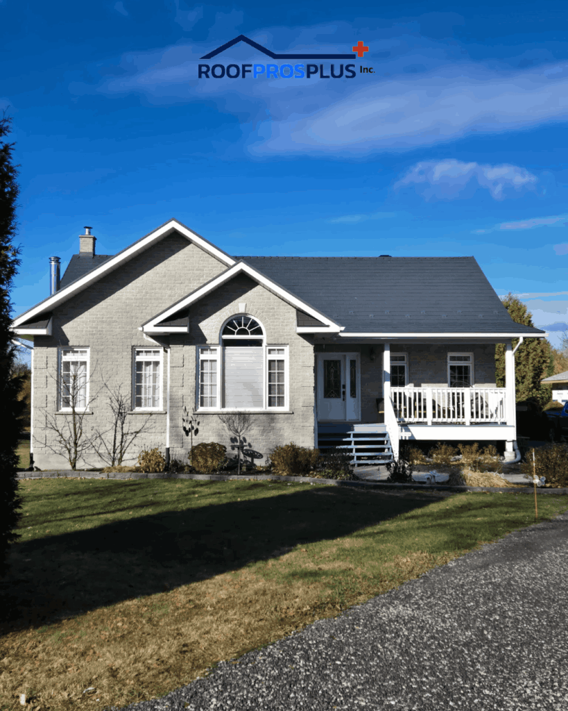 A single-story house with grey brick exterior, white trim, and a front porch. The house sits under a blue sky and a landscaped yard. The logo "Roofpros Plus Inc." is at the top.