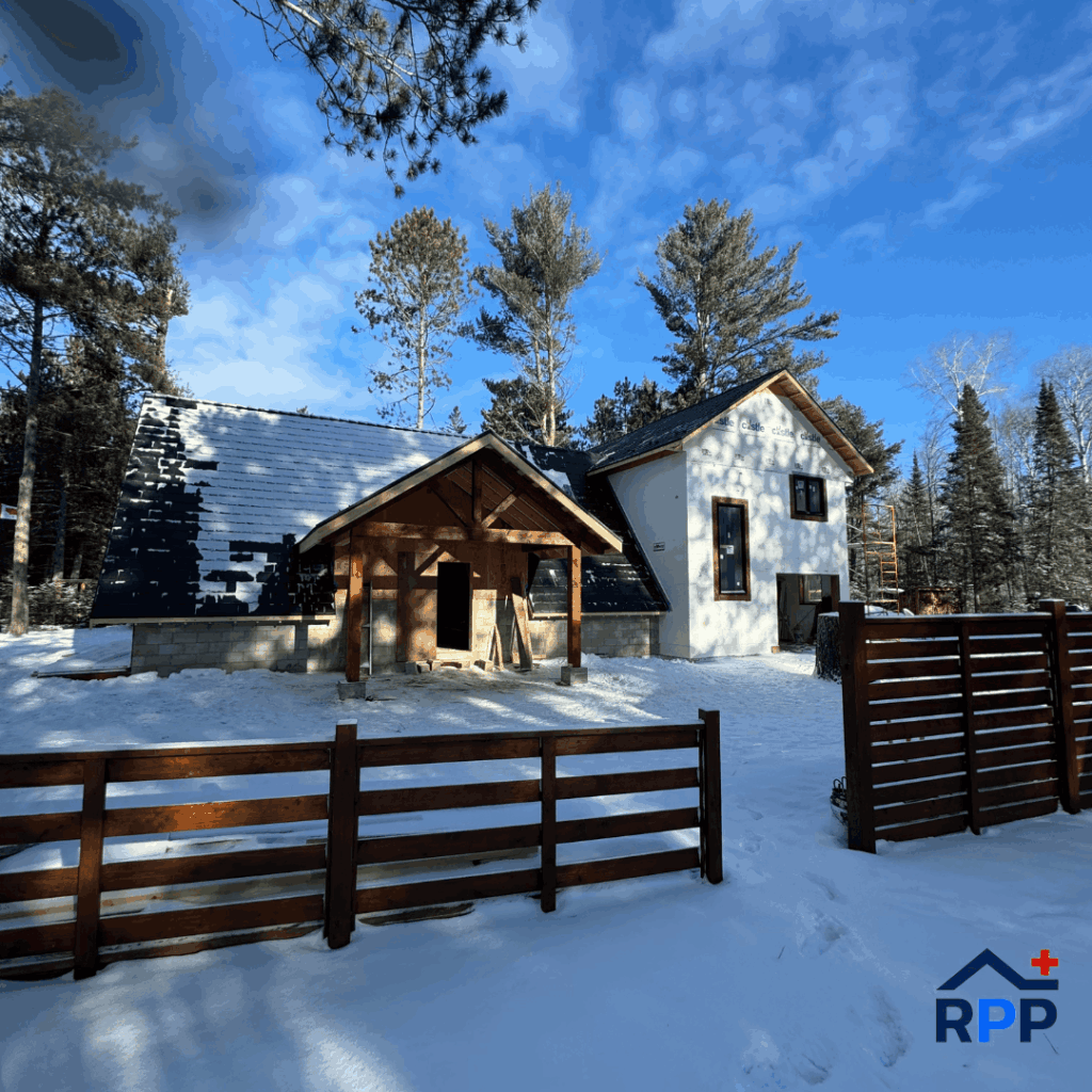 A snow-covered house with wooden accents and dark roofs is surrounded by tall pine trees under a bright blue sky. A wooden fence is in the foreground.