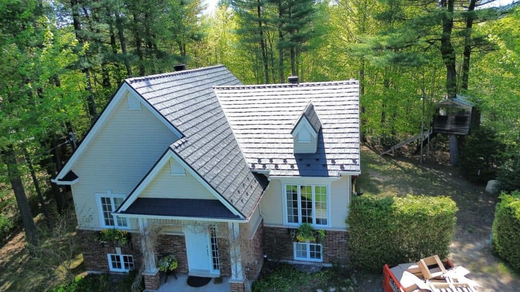 Aerial view of a house with a dark grey metal roof, surrounded by lush green trees and a small playhouse in the background.