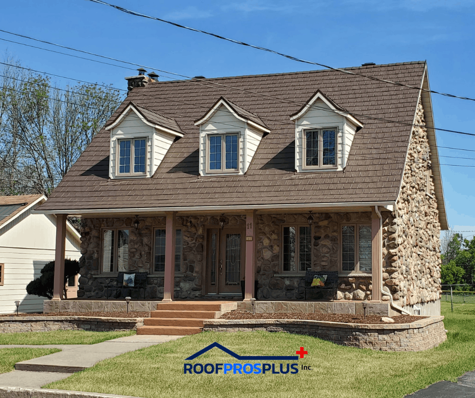 A stone house with a brown metal roof and three dormer windows, surrounded by green lawn. A porch with columns and steps leads to the entrance.