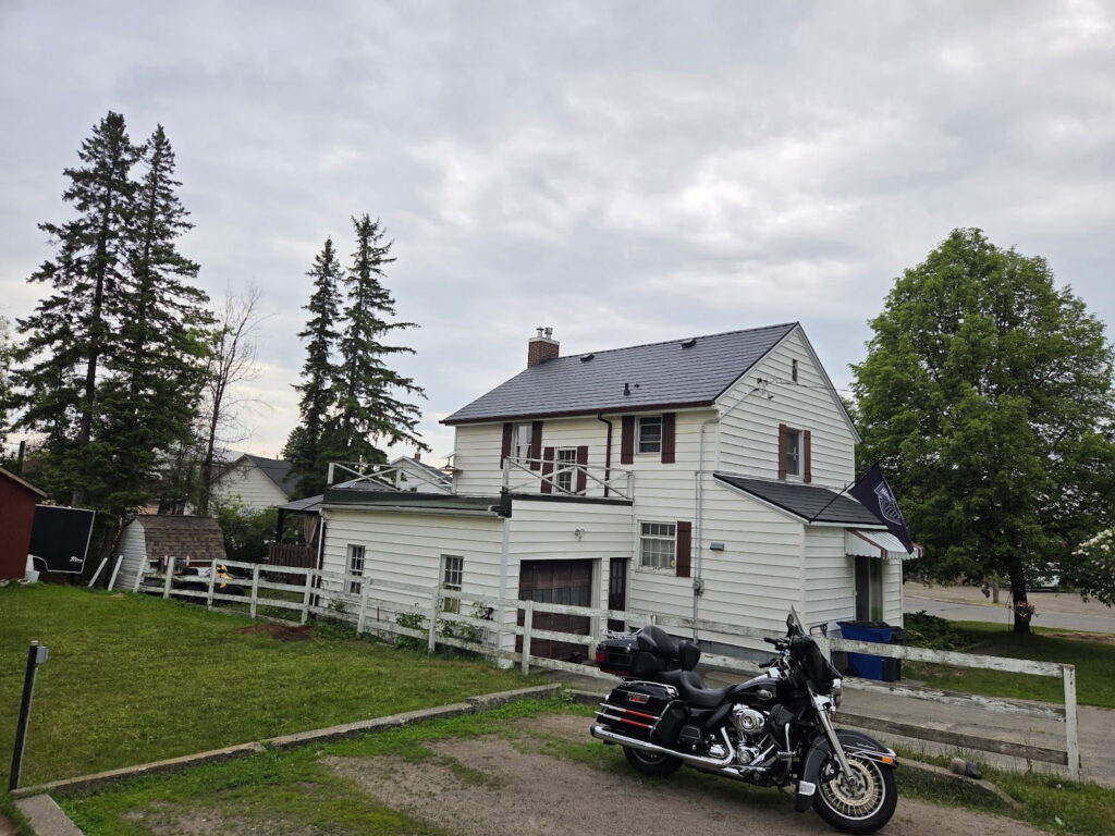 A white two-story house with a sloped metal roof, surrounded by green trees and a fenced lawn. A black motorcycle is parked in the driveway.