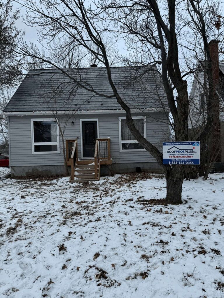 Small grey house with a pitched metal roof and wooden steps, set in a snowy yard. A "Roof Pros Plus" sign is displayed at the front of the house, indicating recent roofing work.